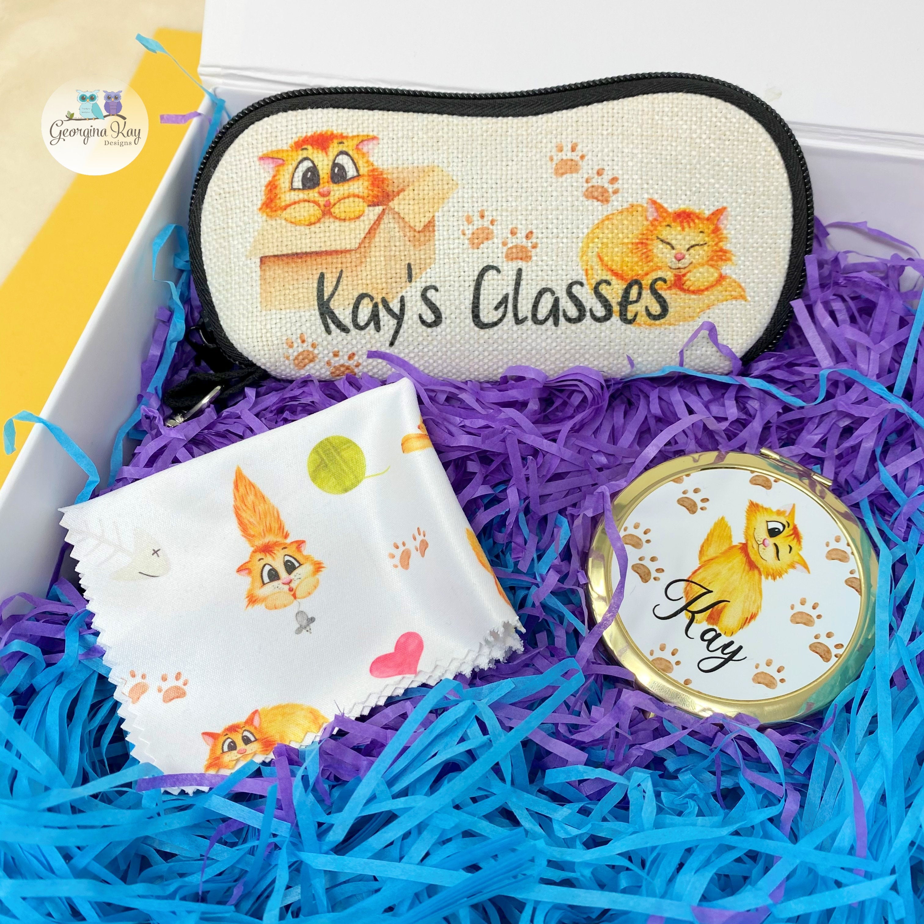 Cat themed glasses case, glass cleaning cloth and pocket mirror. All with a cute ginger cat design in a gift box. Pictured on shredded paper in purple and turquoise. 
