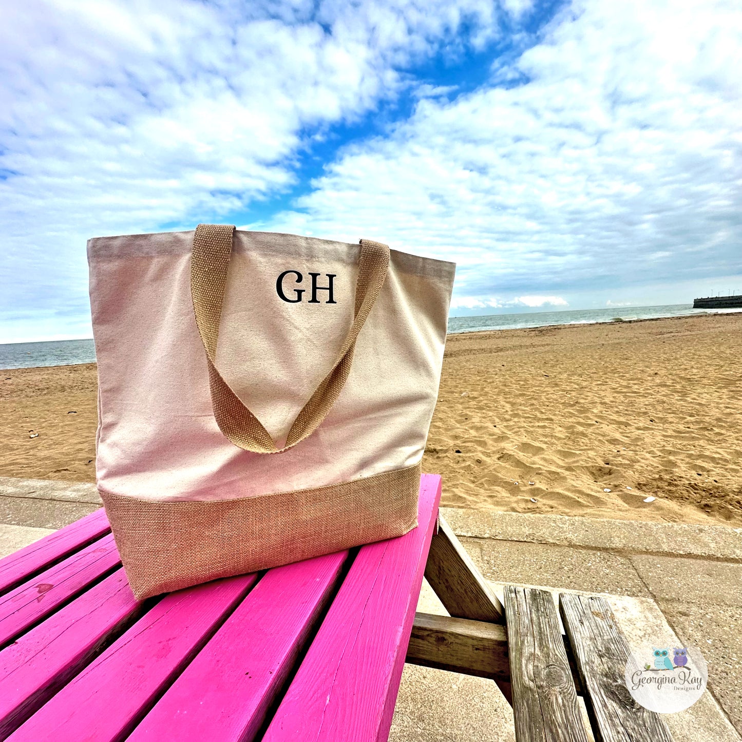 Beige tote bag with 'GH' monogram on a pink table at the beach