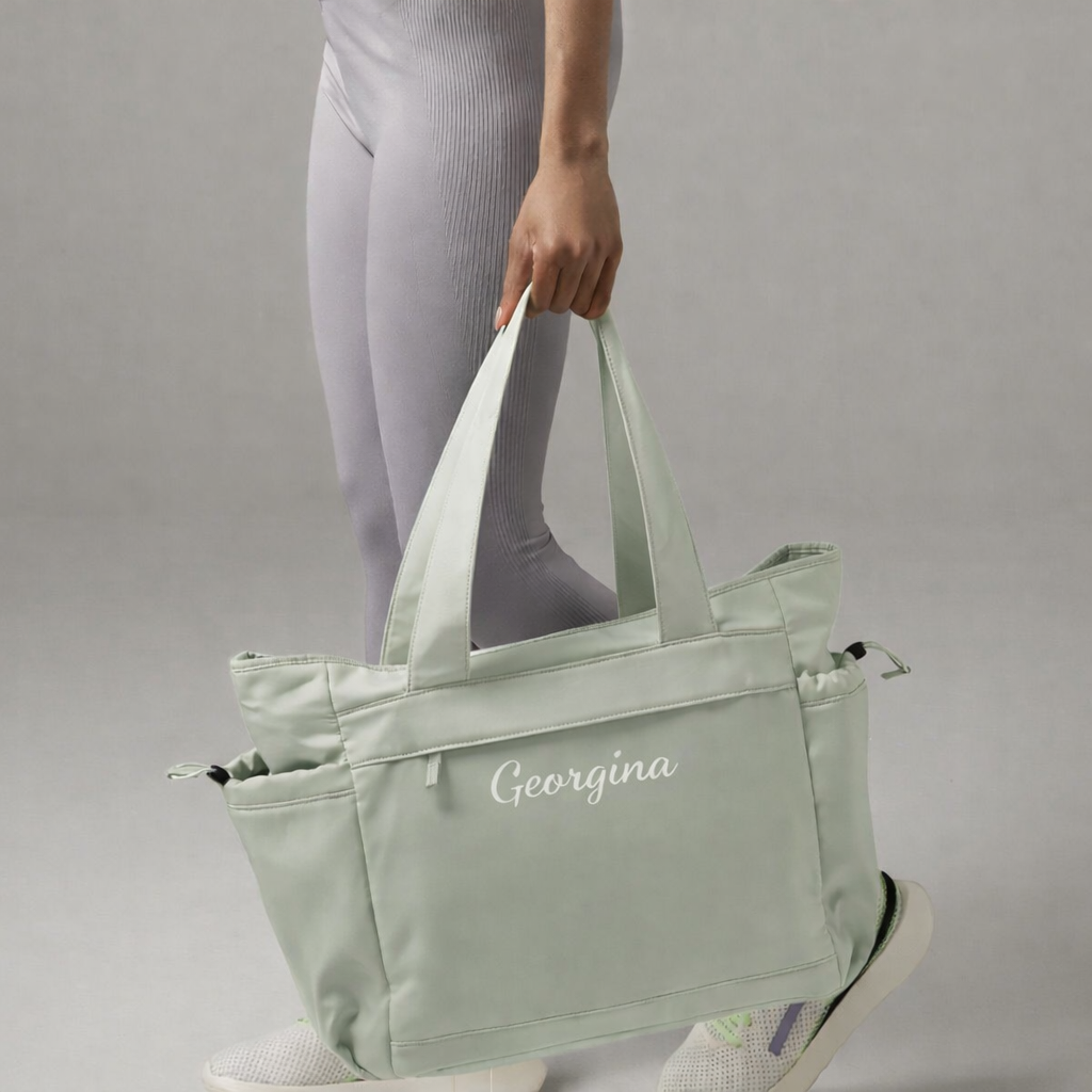 Person holding a light green tote bag with 'Georgina' branding on a gray background