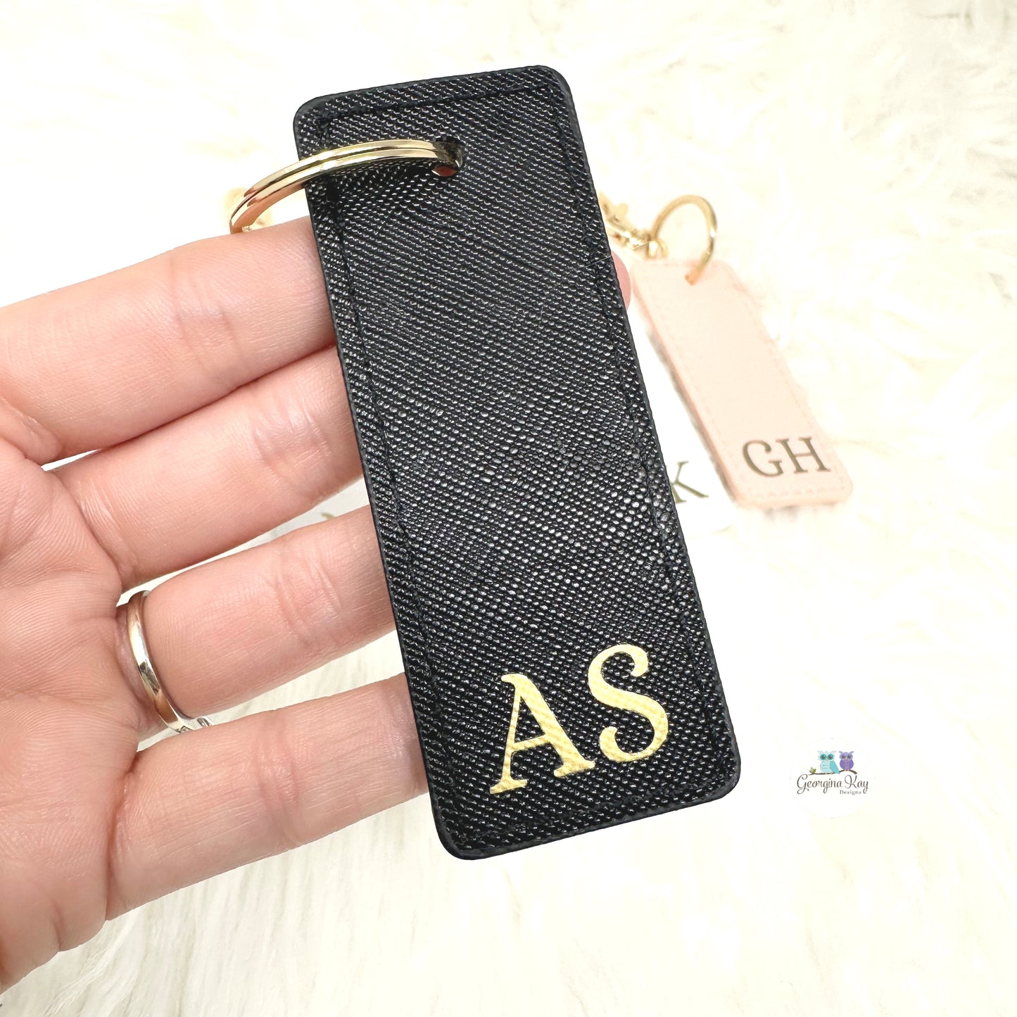 Black keychain with gold initials held in a hand, with a blurred background