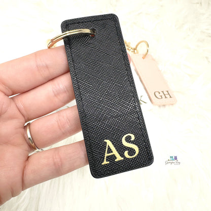 Black keychain with gold initials held in a hand, with a blurred background