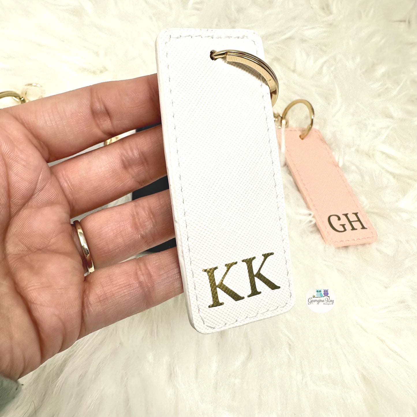 White keychain with gold letters 'KK' held by a hand on a light background