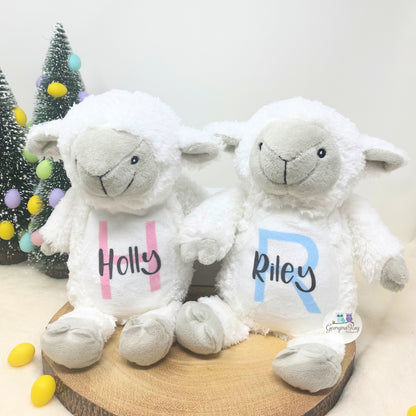 Two white stuffed lambs with 'Holly' and 'Riley' on their chests, sitting on a wooden board with a Christmas tree in the background.