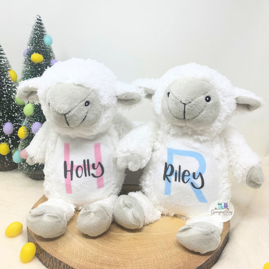 Two white stuffed lambs with 'Holly' and 'Riley' on their chests, sitting on a wooden board with a Christmas tree in the background.