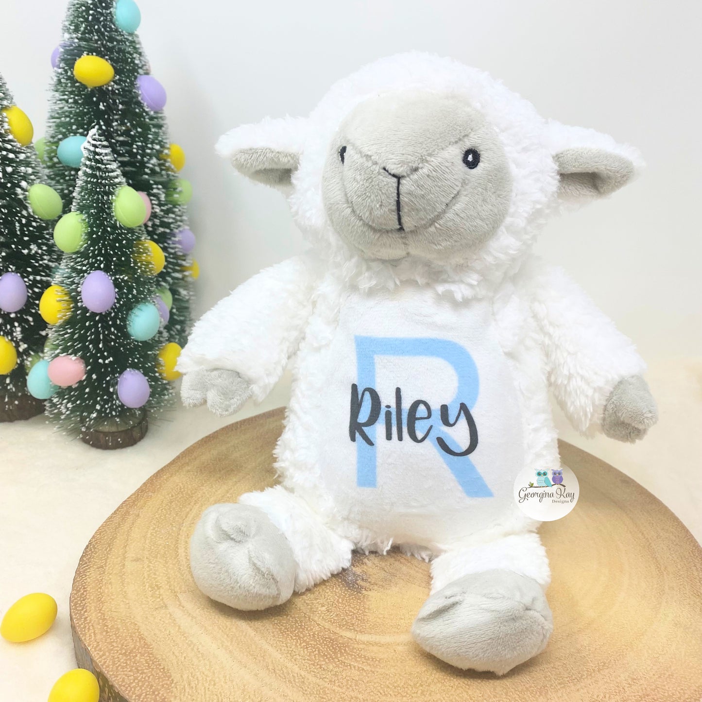 White plush lamb toy with 'Riley' on a wooden surface with Christmas decorations in the background.