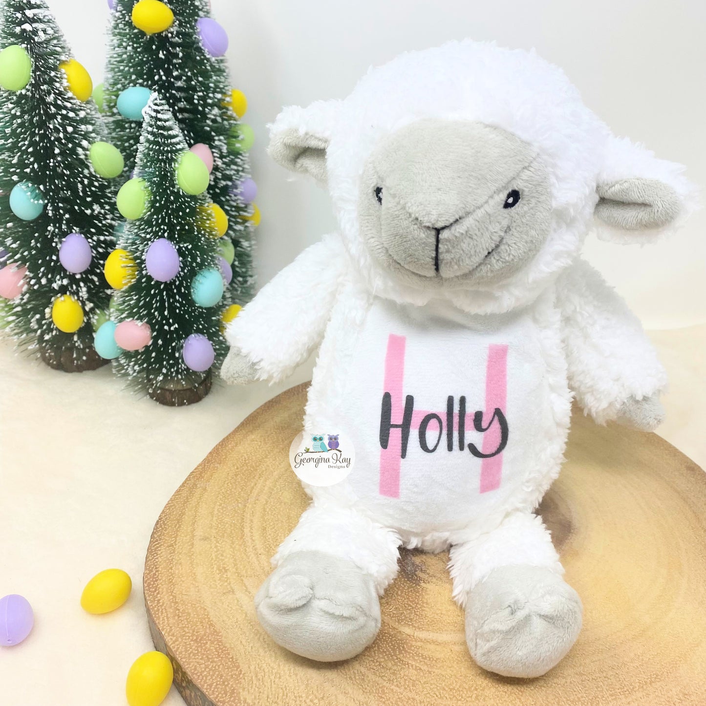 White plush lamb toy with 'Holly' on a wooden stand with decorative trees and eggs in the background.