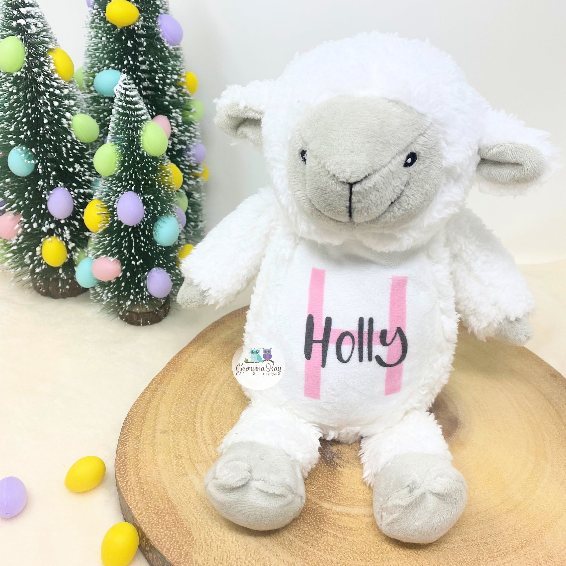 White plush lamb toy with 'Holly' on a wooden stand with decorative trees and eggs in the background.
