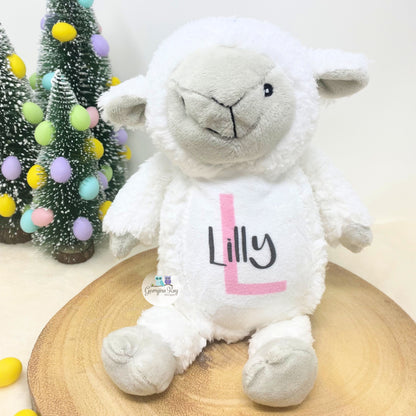 White plush lamb toy with 'Lilly' on a wooden surface with Christmas trees in the background