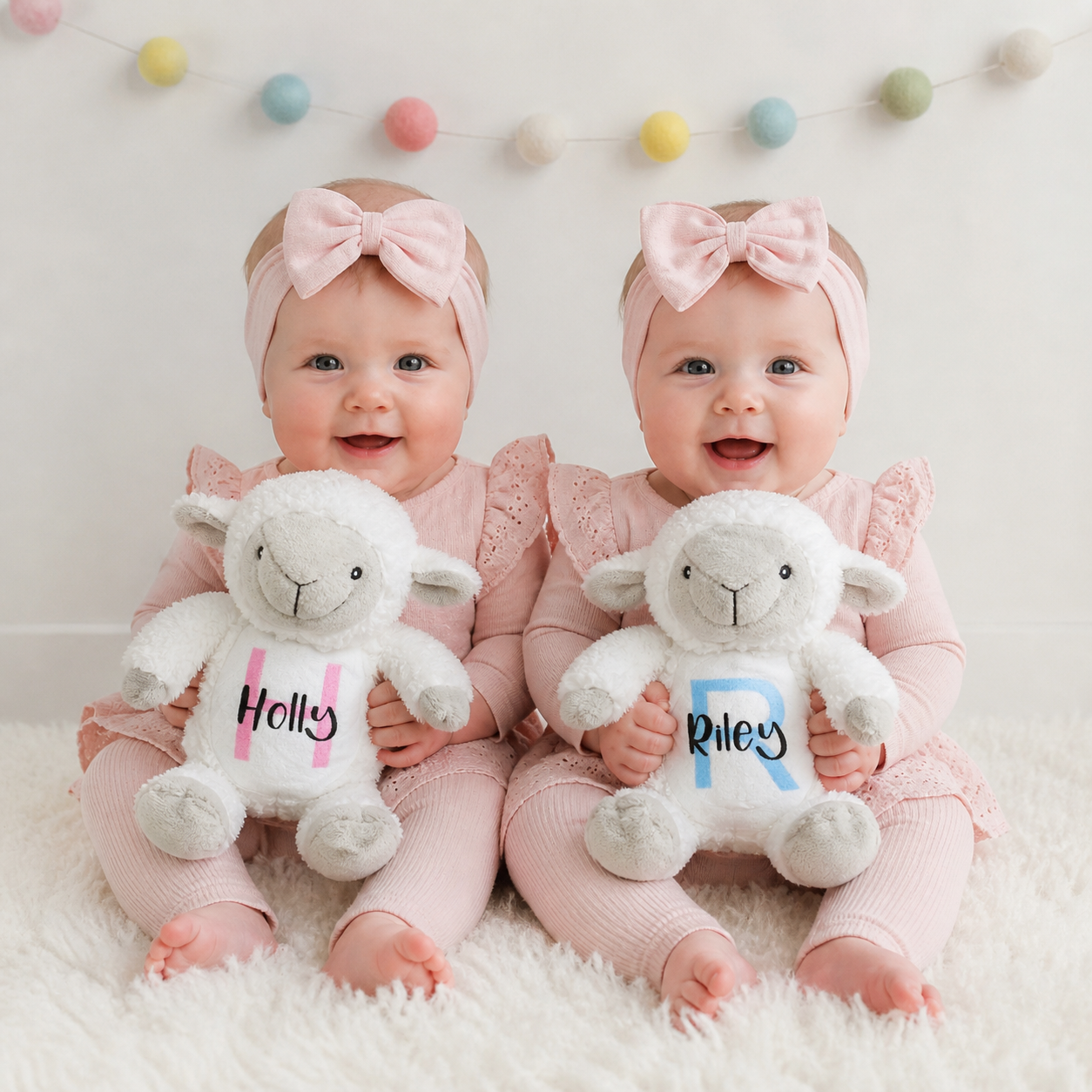 Two babies in pink outfits holding sheep stuffed toys with names on them, sitting on a white surface with colorful balls in the background.