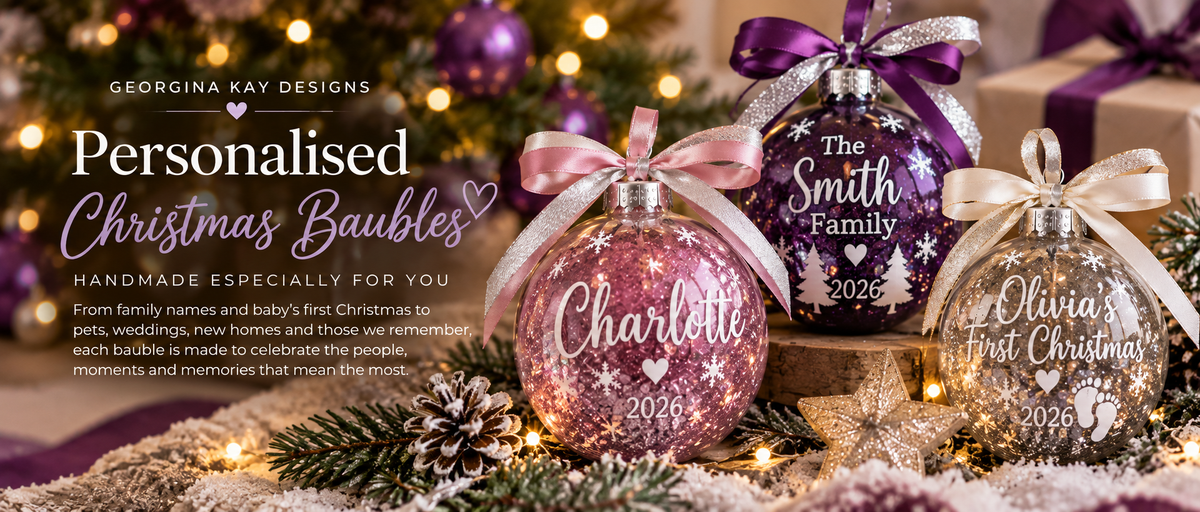 Three personalized Christmas bauble ornaments displayed on a snowy surface with pine branches and warm string lights. The ornaments are pink, purple, and clear/white with silver lettering and bows.