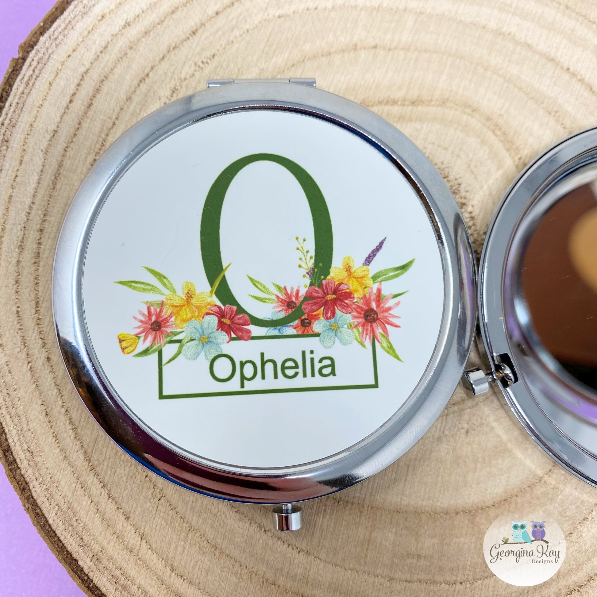 Compact mirror with floral design and 'Ophelia' text on a wooden surface