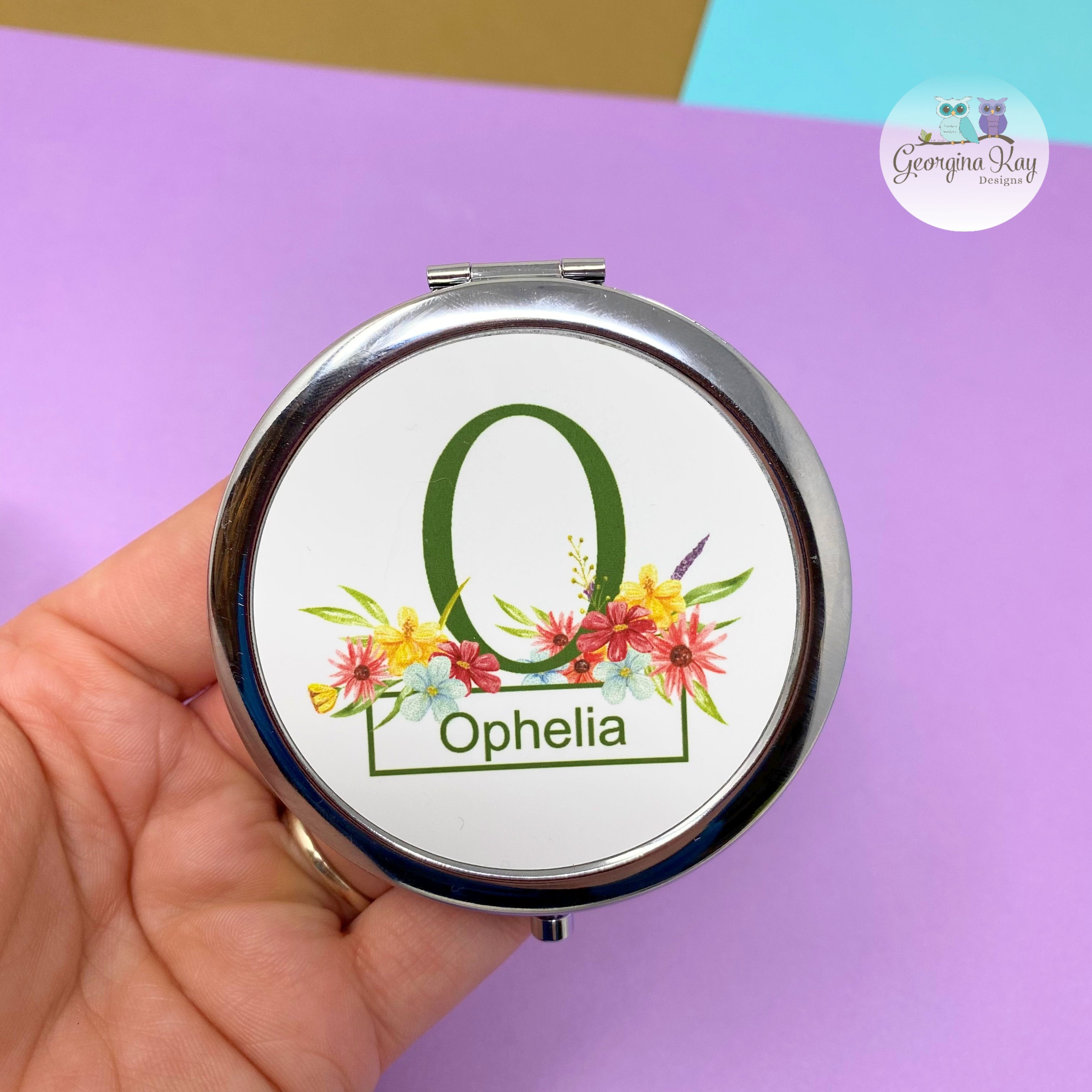 Compact mirror with floral design and 'Ophelia' text held by a hand on a purple background