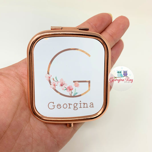 Rose gold compact mirror with floral design and 'Georgina' text held in a hand against a white background.