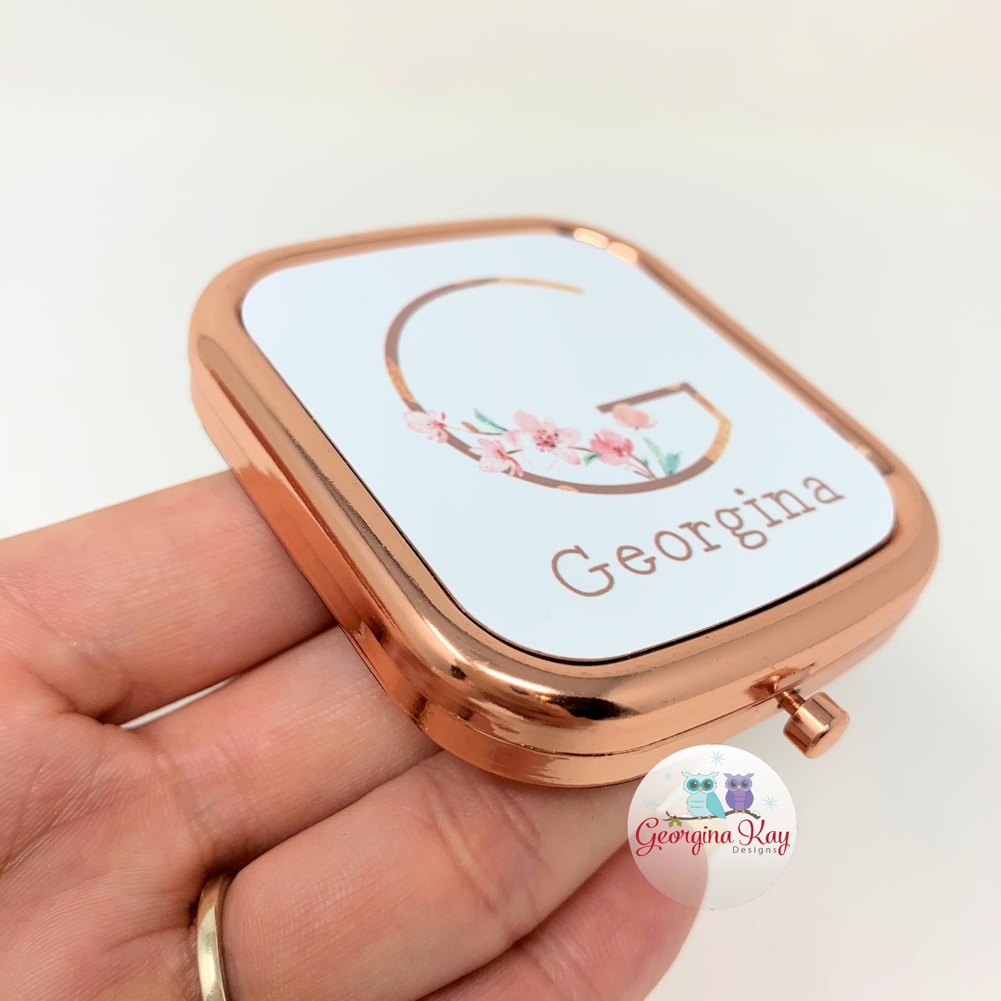 Rose gold compact mirror with 'Georgina' on a light background, held in a hand.