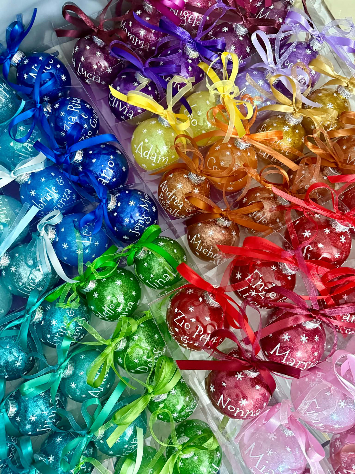 A close-up of multiple clear plastic bags filled with shiny, round Christmas ornaments in a rainbow of colors. The ornaments are wrapped in iridescent foil and tied with colorful ribbons, with white script names like 'Emily', 'Sophia', and 'Mia' printed on them.